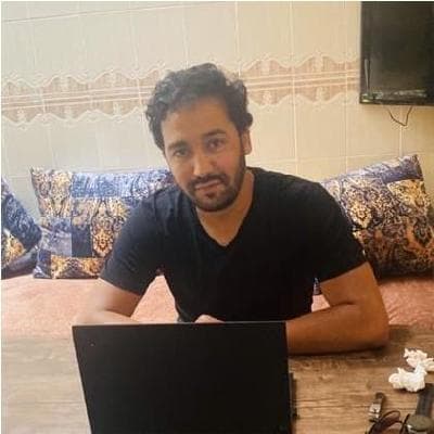 Zakaria ARRAMA, a man with dark curly hair and a short beard, wearing a black v-neck t-shirt, sitting at a wooden table behind a black laptop in a traditional Moroccan living room with patterned blue-and-white cushions on a bench seat behind him.